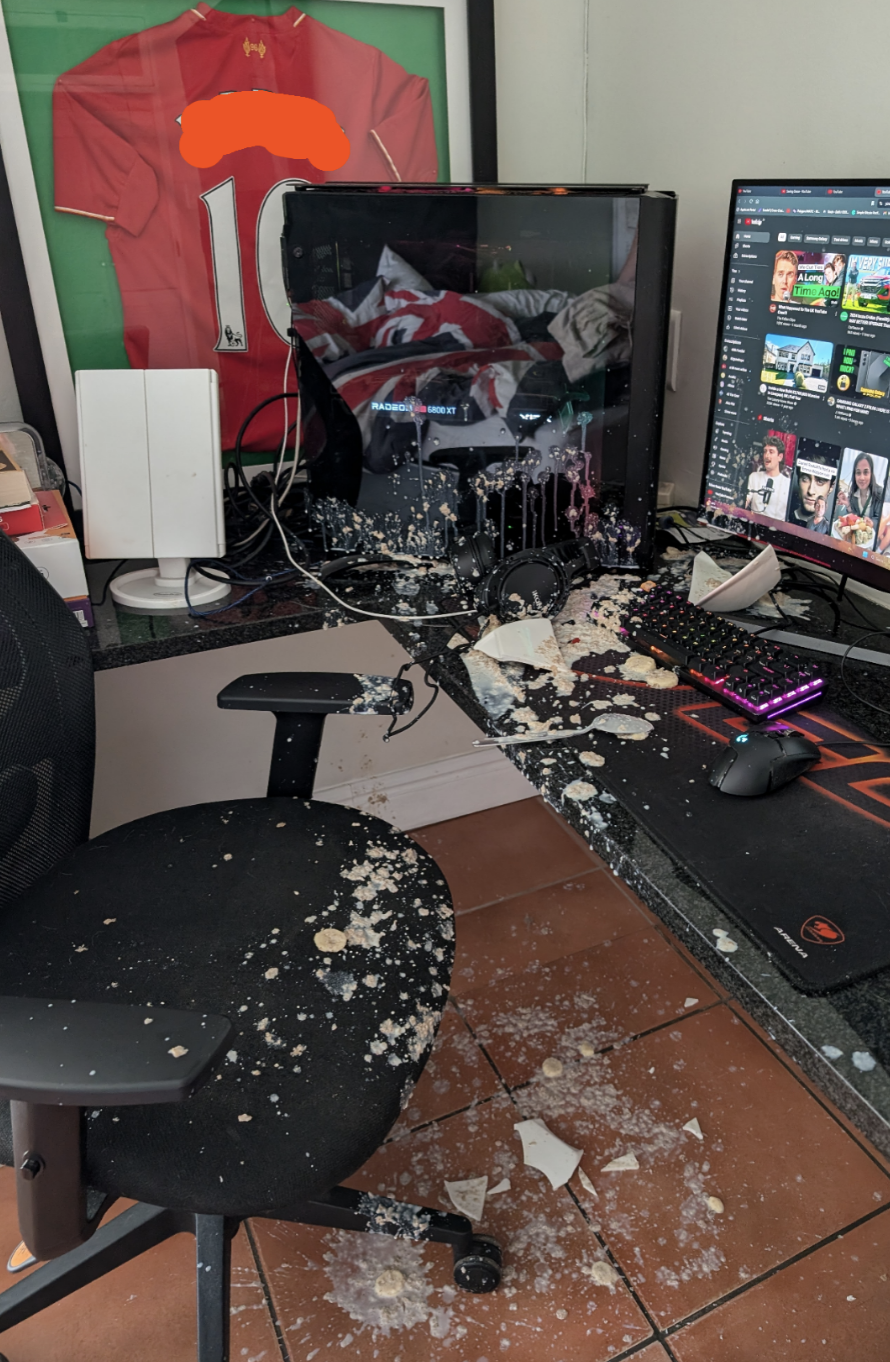A computer desk with a broken bowl and food spilled everywhere, covering the desk, keyboard, mouse, and chair. A framed sports jersey and a monitor are in the background