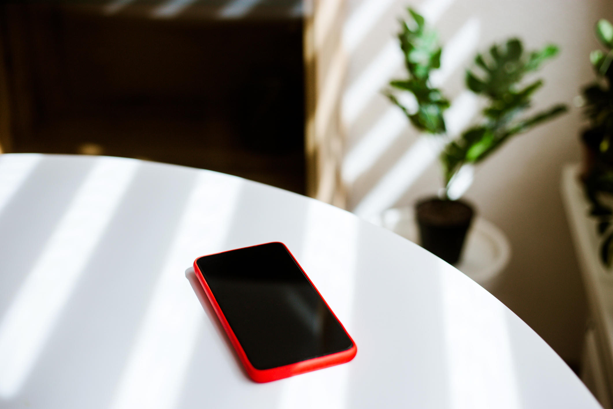 A red smartphone lies face up on a white table, with two potted plants in the background