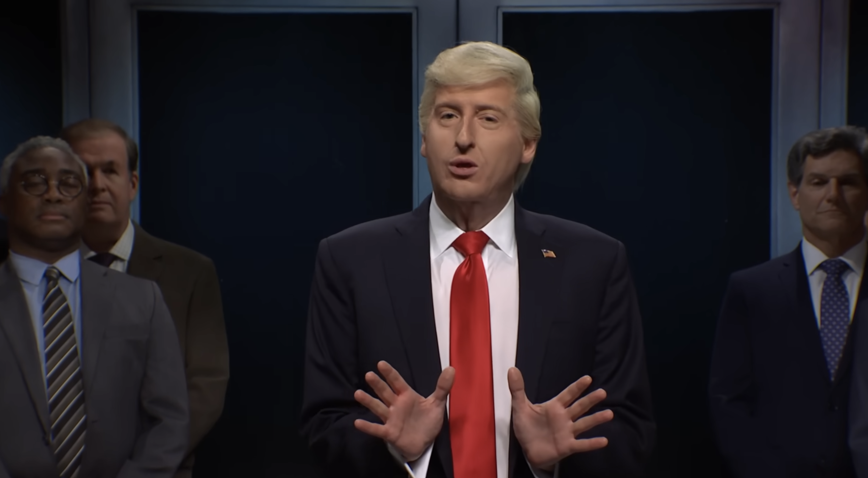 A man resembling Donald Trump speaks while gesturing with his hands, with four other men standing behind him in suits and ties