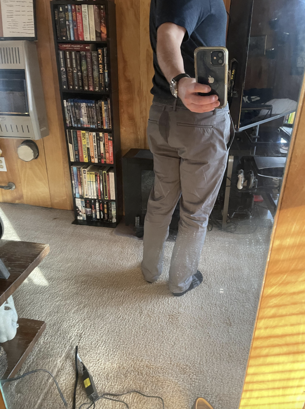 A person stands in front of a mirror, holding a phone to take a photo of their back and gray pants. There is a noticeable stain on the pants. Bookshelves and a heater are in the background