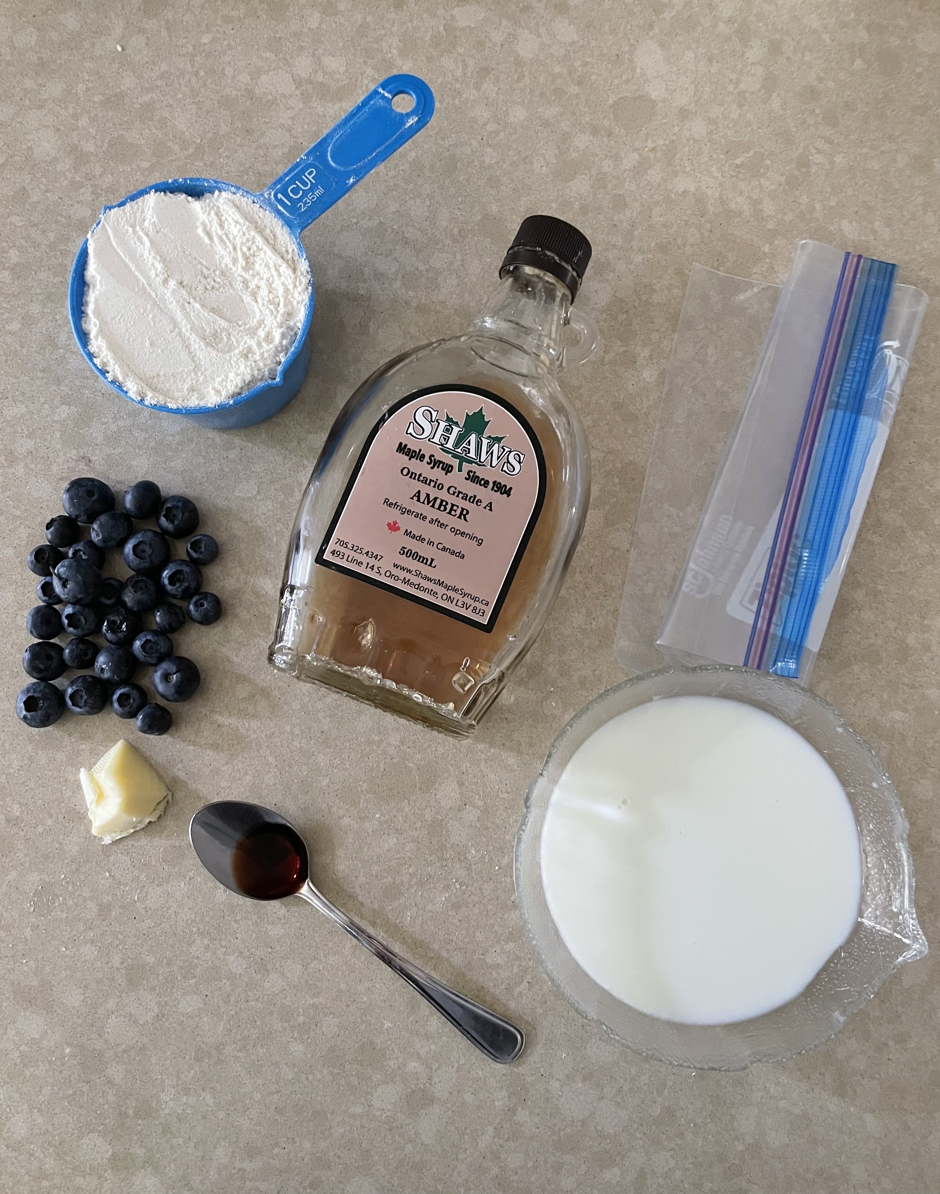 Ingredients for making pancakes: flour, blueberries, butter, syrup, vanilla extract, milk, and a plastic bag