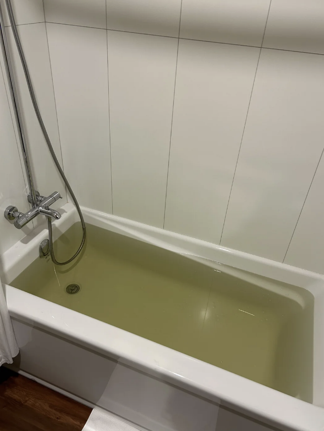 A bathtub with yellowish water, a shower hose, and white tiled walls