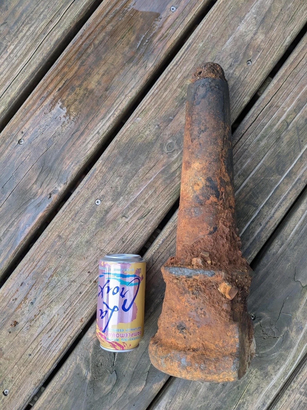 A rusty, old metal object lies on a wooden surface next to a LaCroix can for scale comparison