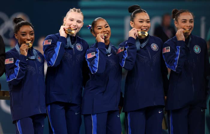 Simone Biles, Hezly Rivera, Jordan Chiles, Sunisa Lee, and Jade Carey in athletic tracksuits, holding gold medals and posing while biting them