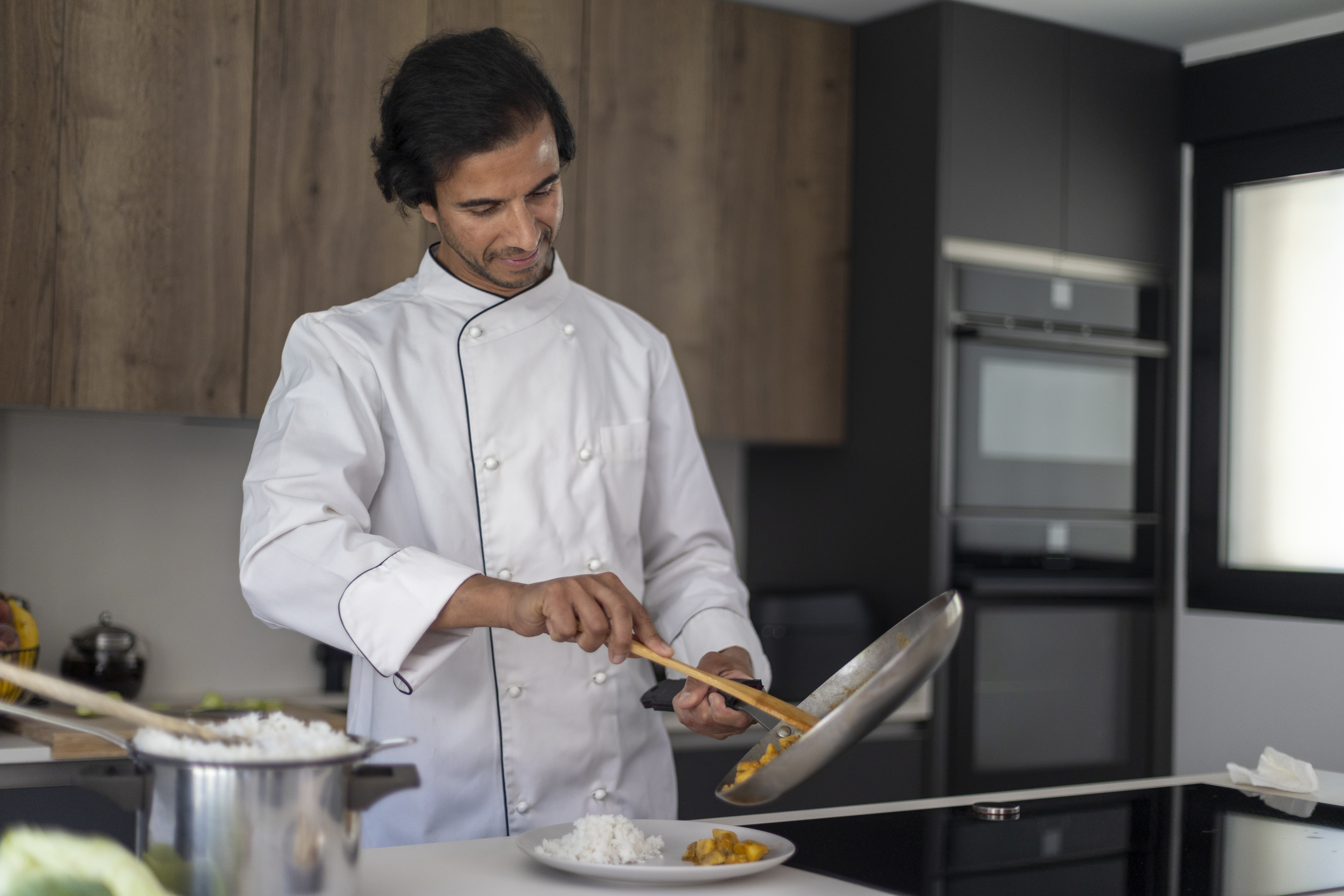 A chef, wearing a white double-breasted chef's coat, is plating food from a frying pan in a modern kitchen with wooden cabinets. No other persons are in the image