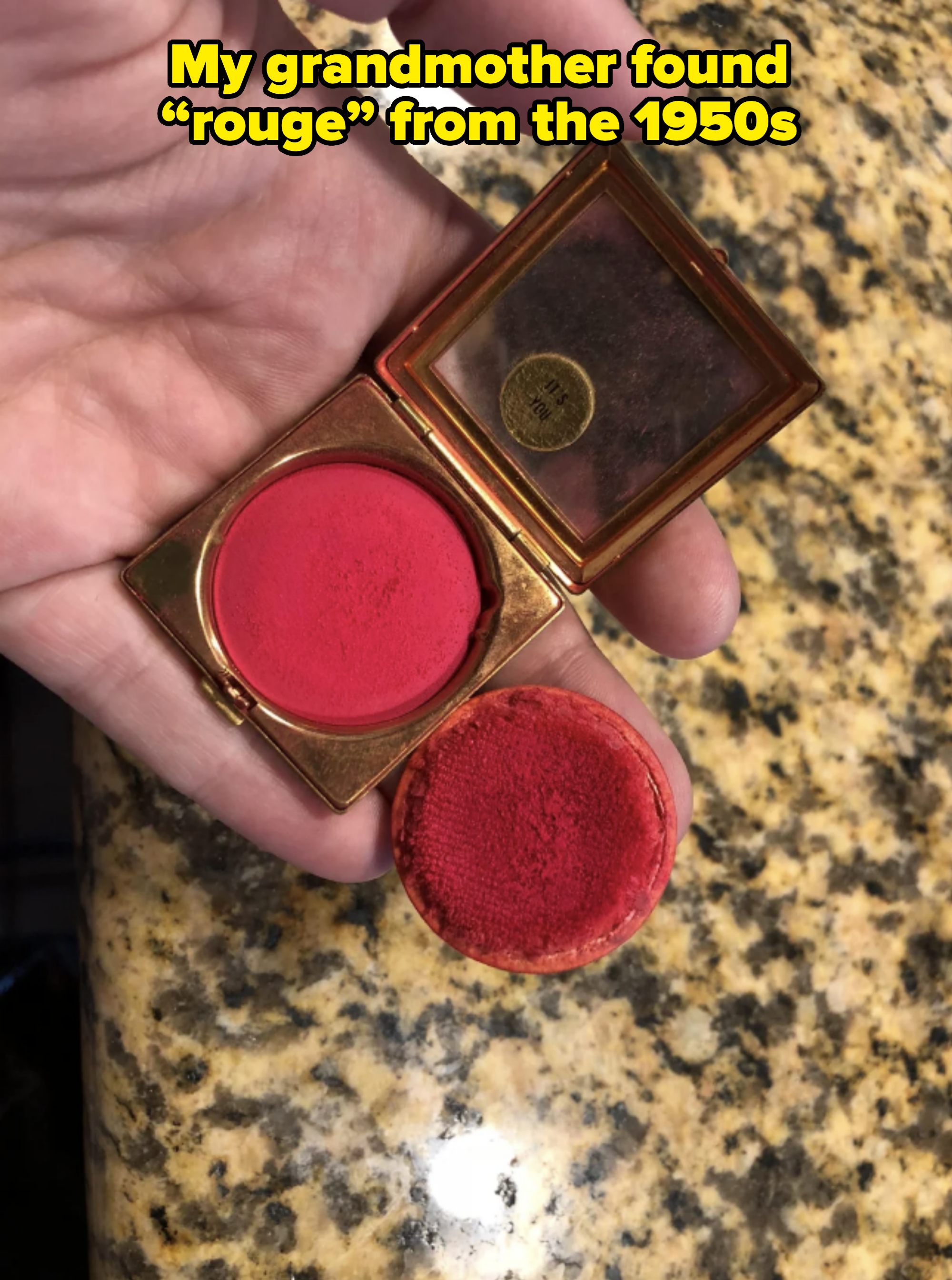 A hand holding a small, open compact with a mirror and red blush inside, over a granite countertop