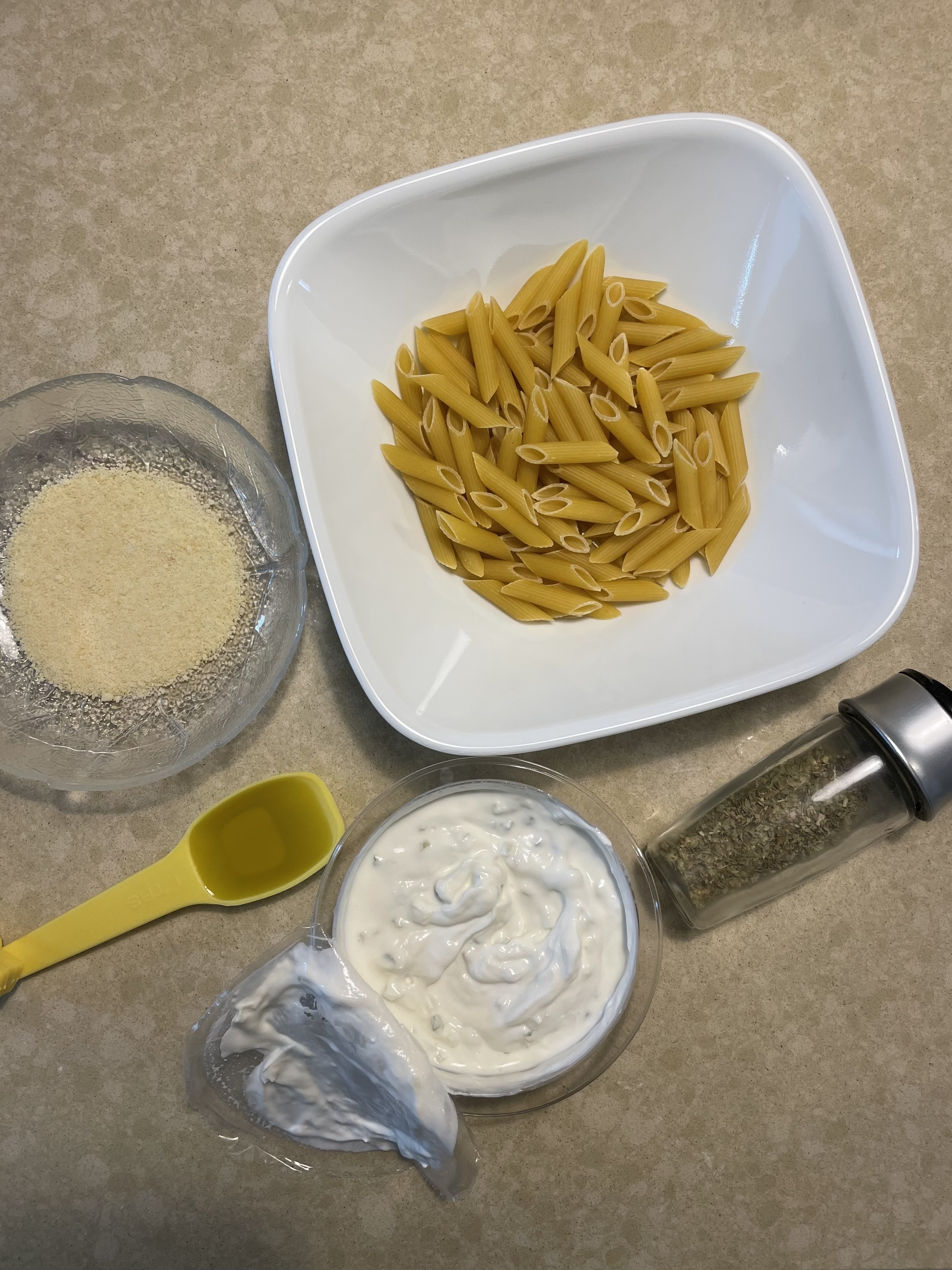 Penne pasta, grated cheese, olive oil in a yellow spoon, Italian seasoning, and a creamy white sauce displayed on a kitchen countertop
