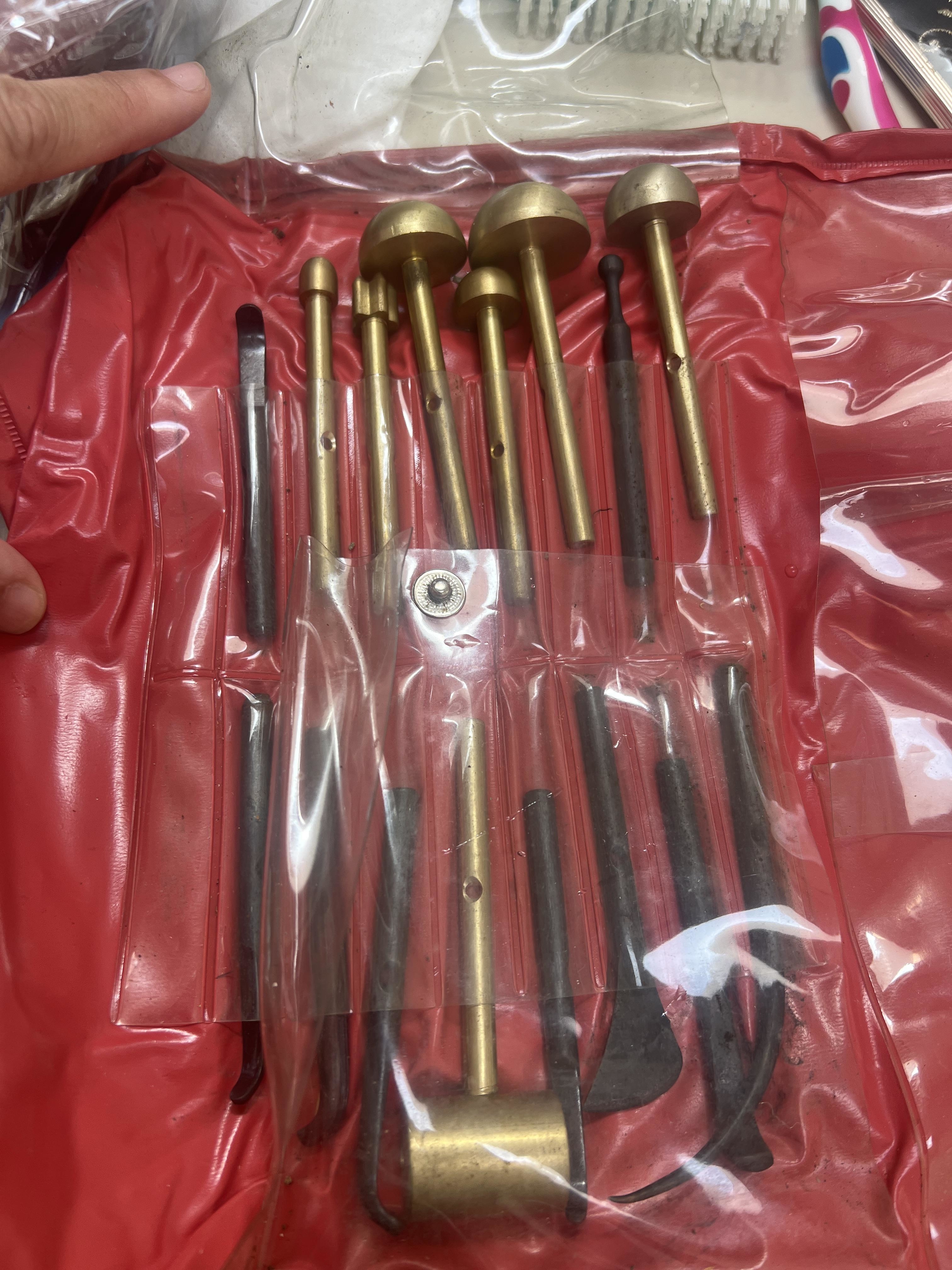A set of manual brass tools and metal implements laid out in a pinkish red organizer pouch