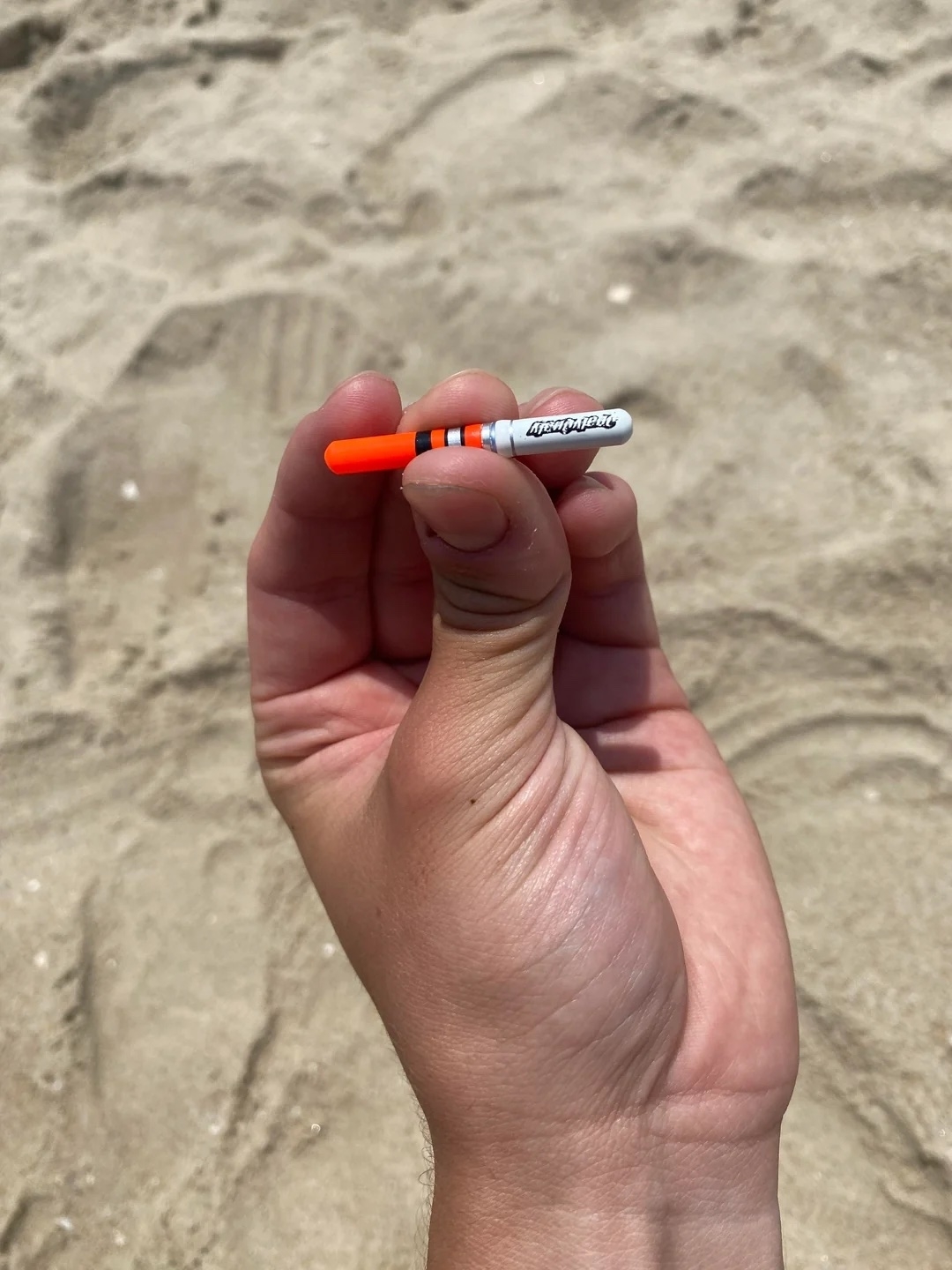 A hand holding a small orange and white cylindrical capsule on a sandy beach