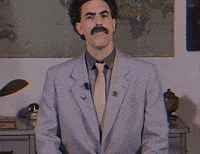 Sacha Baron Cohen, dressed as Borat in a grey suit with a beige tie, stands in front of a world map, looking straight at the camera