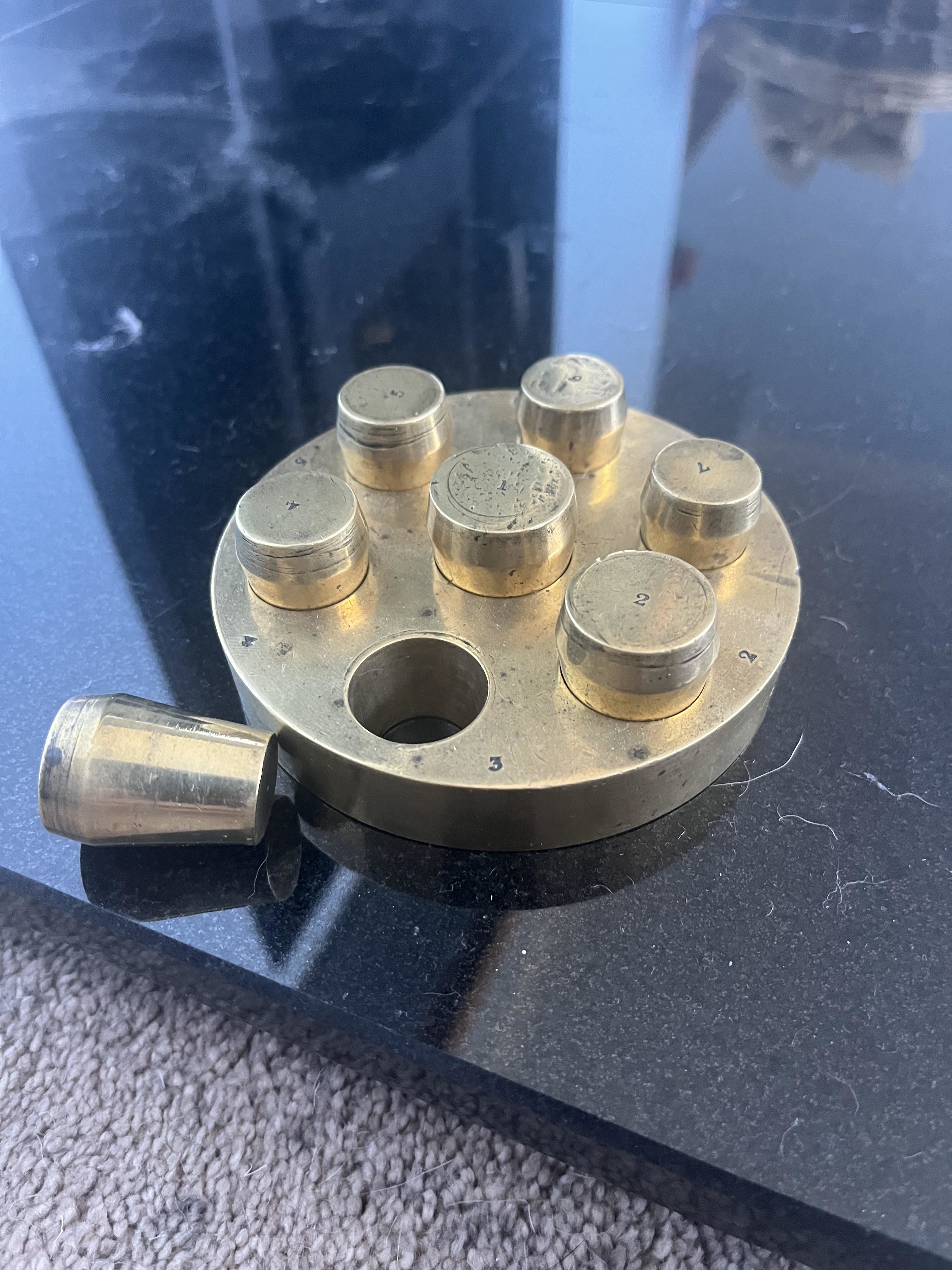 A large, brass revolver-shaped cylinder with six chambers, showing one empty chamber and the extra cylinder nearby