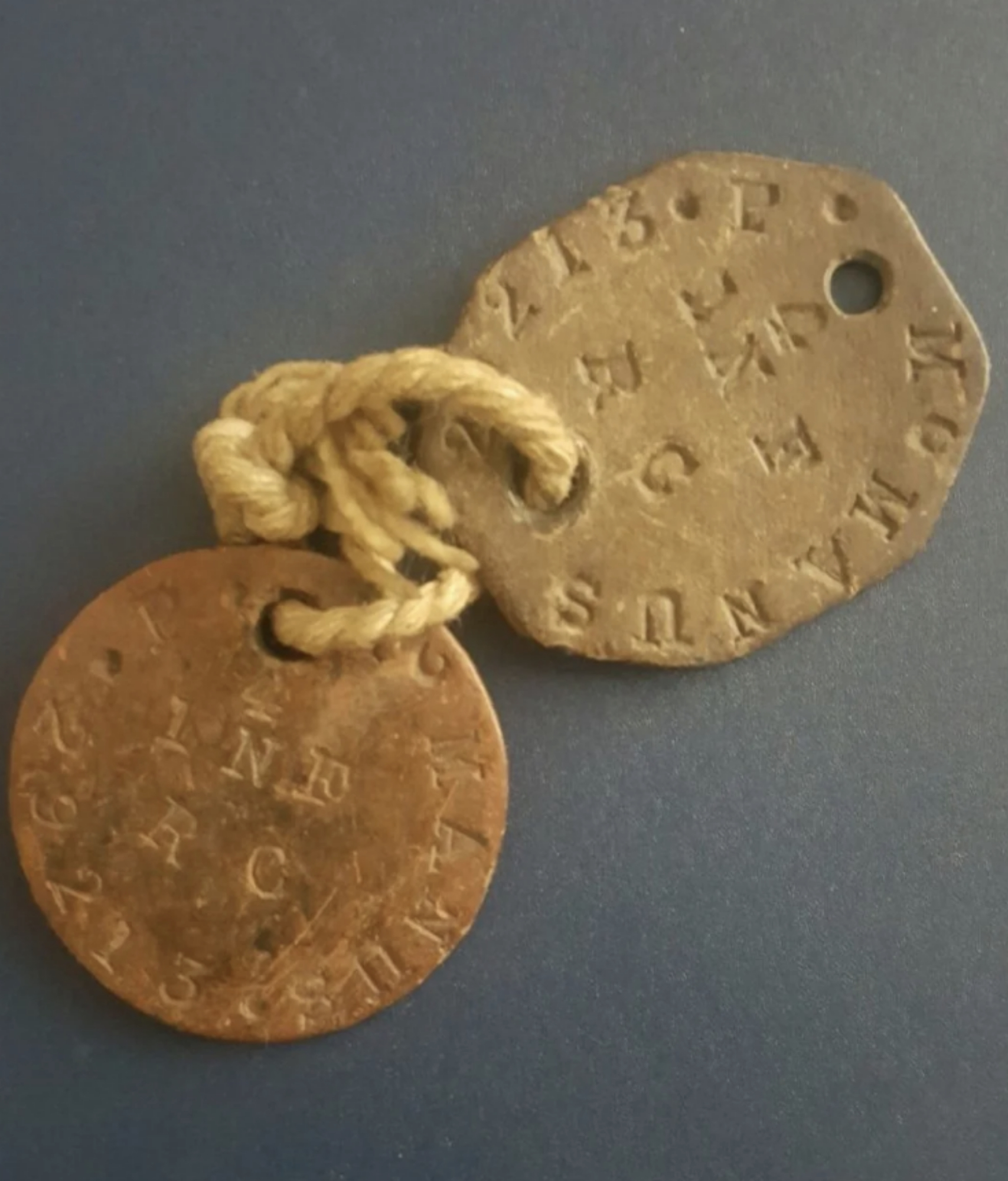Two old metal tags attached by a rope, one with a hexagonal shape and the other round, each engraved with letters and numbers