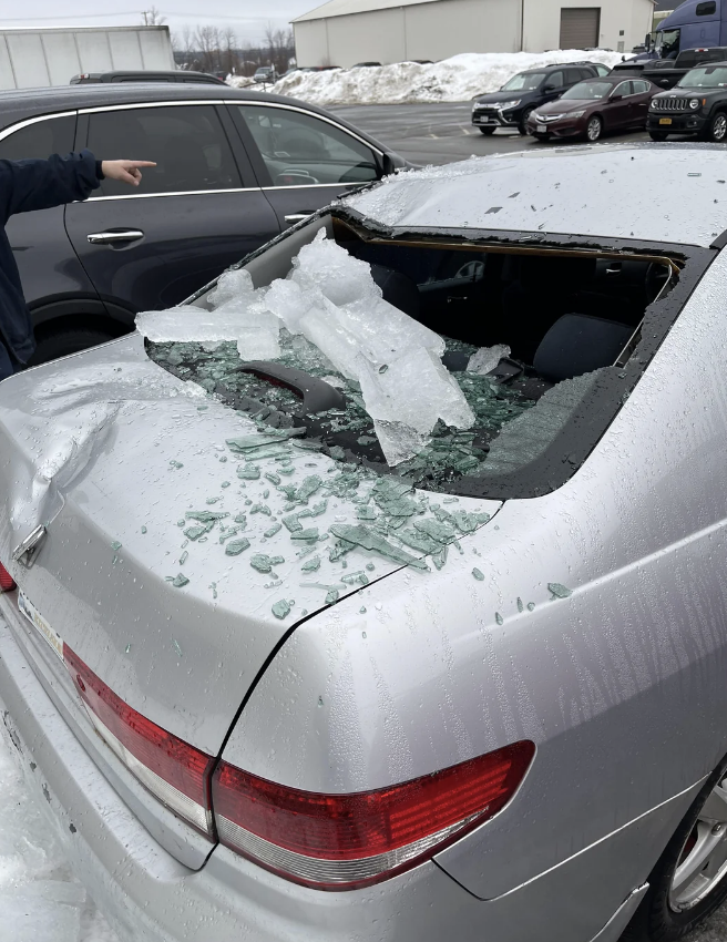 A parked car with its rear windshield shattered, covered in ice and broken glass, while a person points towards it. Snow and other vehicles are visible in the background