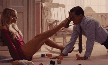 Margot Robbie in a red bodysuit playfully pushes Leonardo DiCaprio's face with her foot while he is on the floor in a child's room with toys