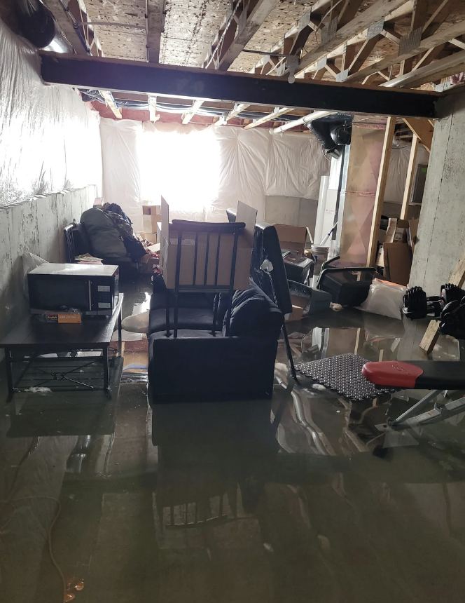 A flooded basement with various furniture, boxes, and equipment partially submerged in water. The light from a window is visible in the background