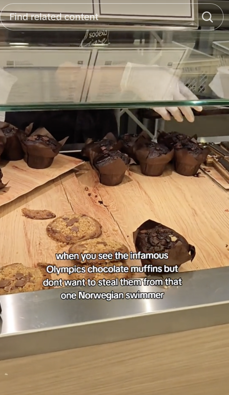 A bakery display case with chocolate muffins and cookies. Text: "when you see the infamous Olympics chocolate muffins but don't want to steal them from that one Norwegian swimmer."