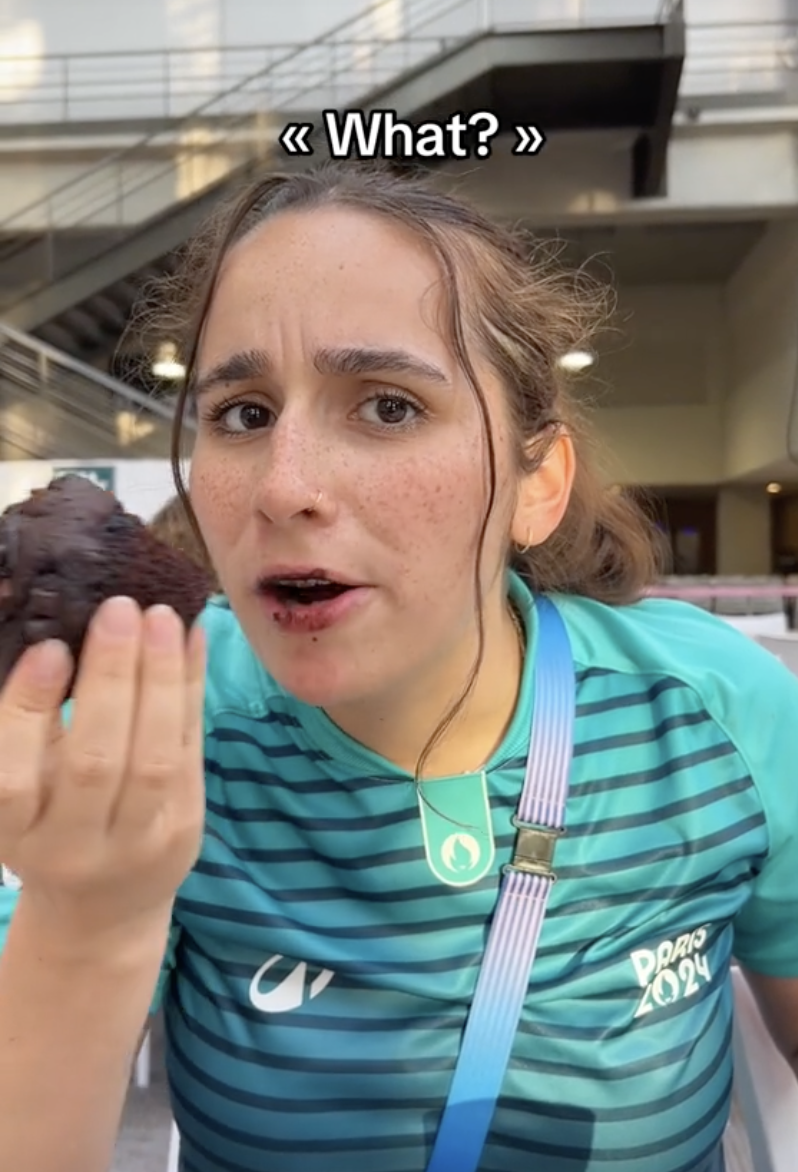 A woman in a green shirt labeled "Paris 2024" holds a chocolate dessert to her mouth with the text "What?" above her