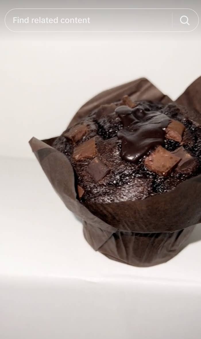 Close-up of a chocolate muffin topped with chunks and a drizzle of chocolate sauce, placed in a brown paper wrapper