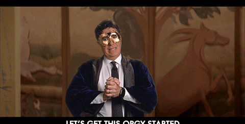 Stephen Colbert in a suit and robe, wearing a gold mask, with &quot;LET'S GET THIS ORGY STARTED&quot; text at the bottom