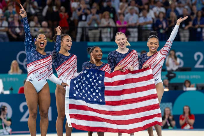 Simone Biles, Jordan Chiles, Grace McCallum, MyKayla Skinner, and Sunisa Lee celebrate holding a U.S. flag at a gymnastics event