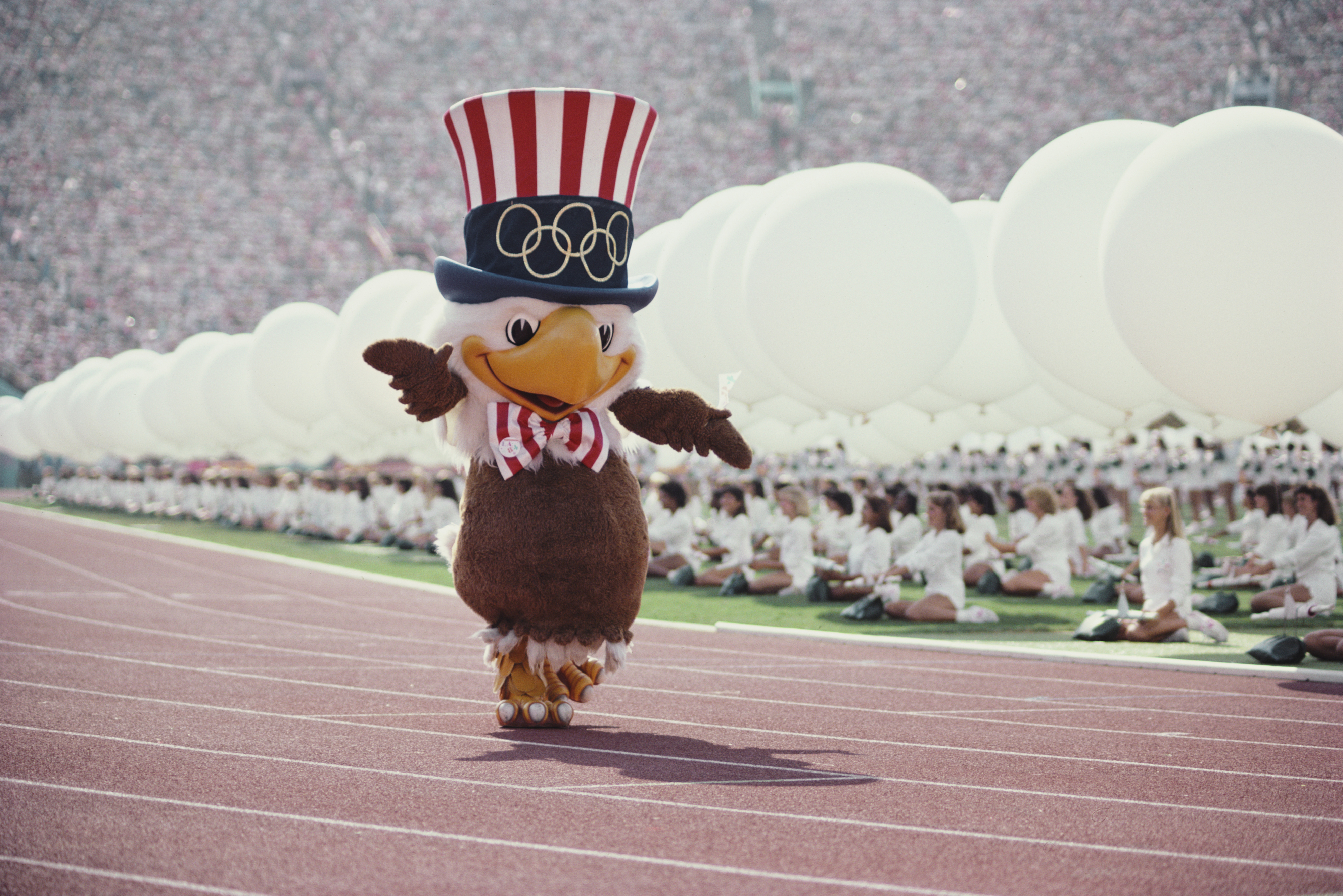 Olympic mascot Sam the Eagle, wearing a top hat with the Olympic rings, walks on a track with a crowd of people and large balloons in the background