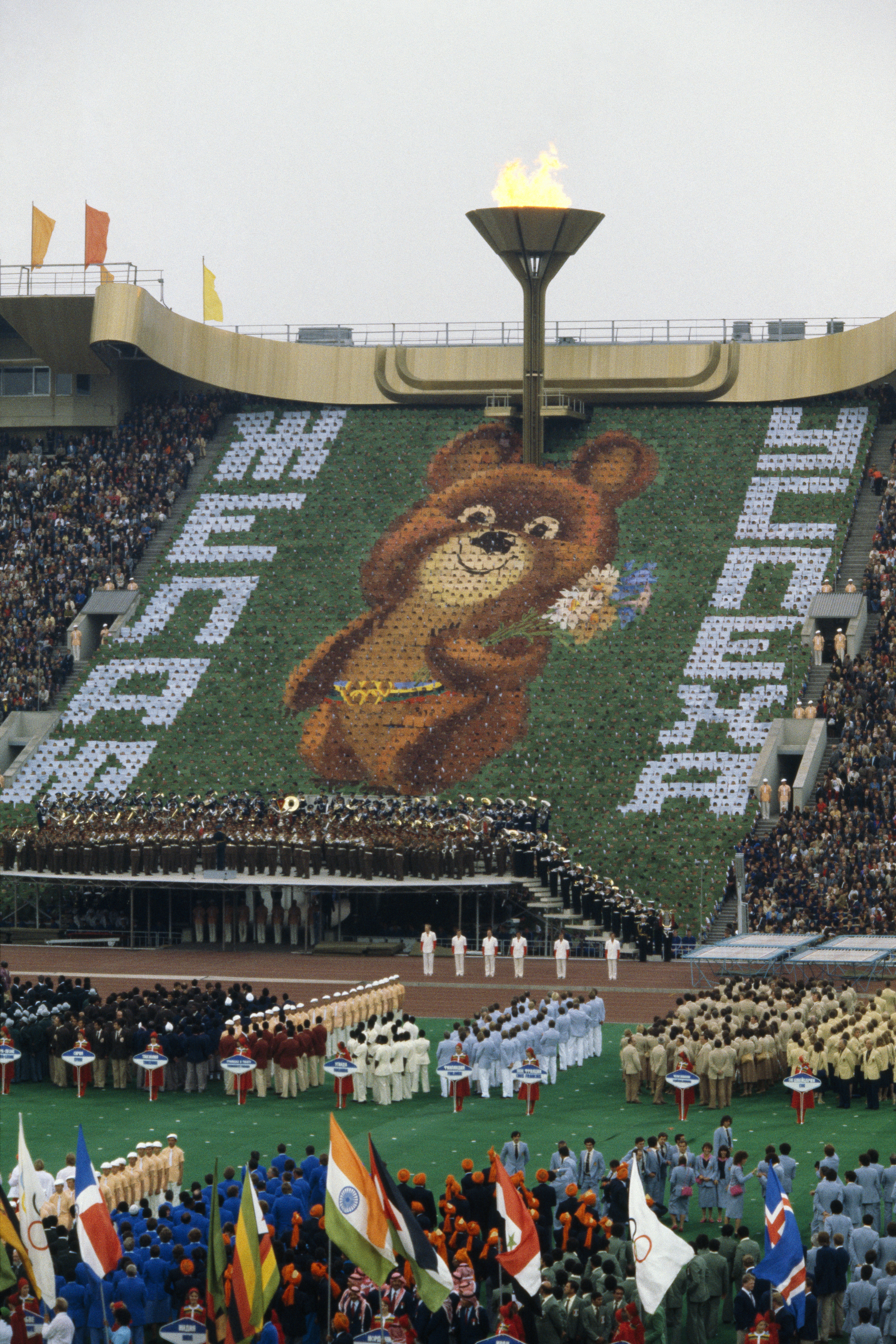 A large crowd at a stadium for the 1980 Moscow Olympics opening ceremony. A giant mosaic of Misha the bear is displayed under the Olympic torch