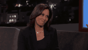 Kamala Harris sitting on a talk show couch, smiling and shrugging. Background shows a night cityscape