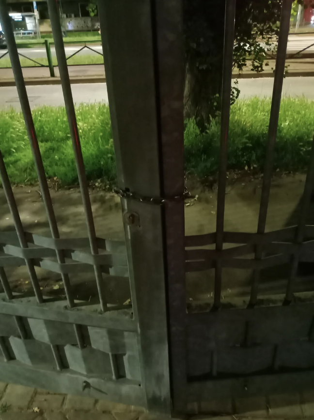 A gate chained and locked, partially open, with a tree and sidewalk in the background