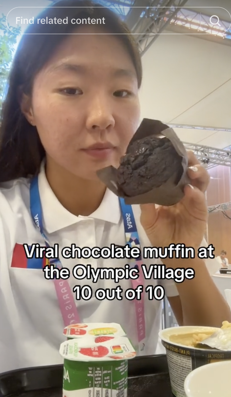 Person eats a chocolate muffin at the Olympic Village, caption reads, "Viral chocolate muffin at the Olympic Village 10 out of 10."
