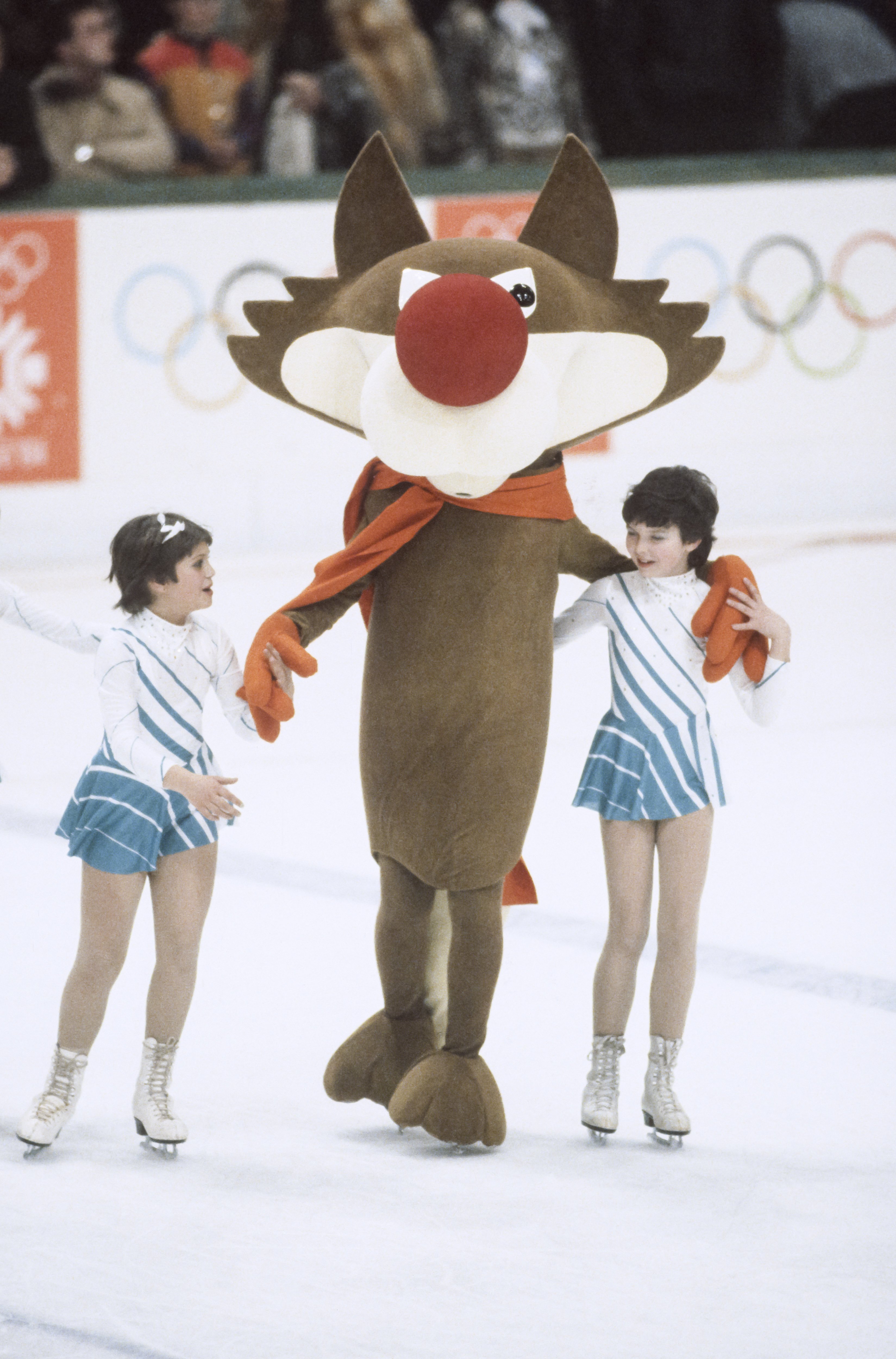 Two young figure skaters hold hands with an Olympic mascot. The skaters are dressed in matching outfits with ice skates. Olympic rings are visible in the background