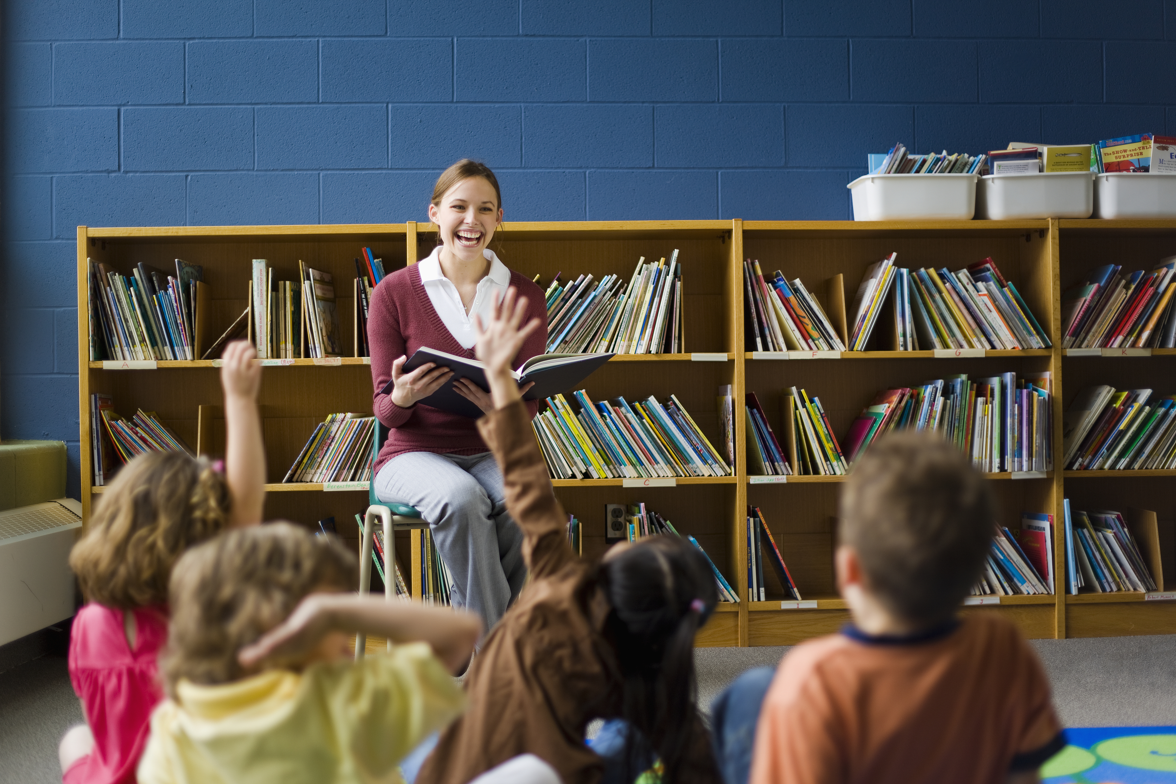 A teacher is sitting on a stool, reading to a group of young children who are sitting on the floor with their hands raised in a classroom library