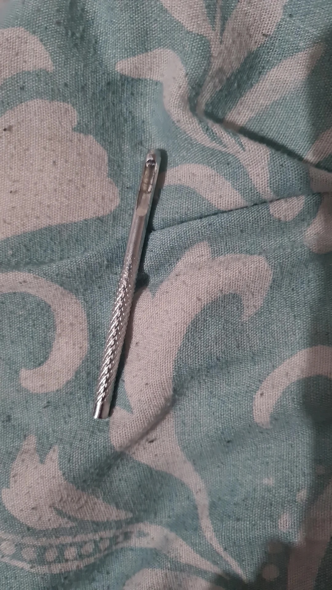 A small, slender, metal object with a textured handle is lying on a fabric with an intricate pattern