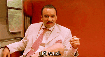 A man in a suit is sitting on a train, holding a cigarette, and saying, &quot;Mi scusi.&quot;
