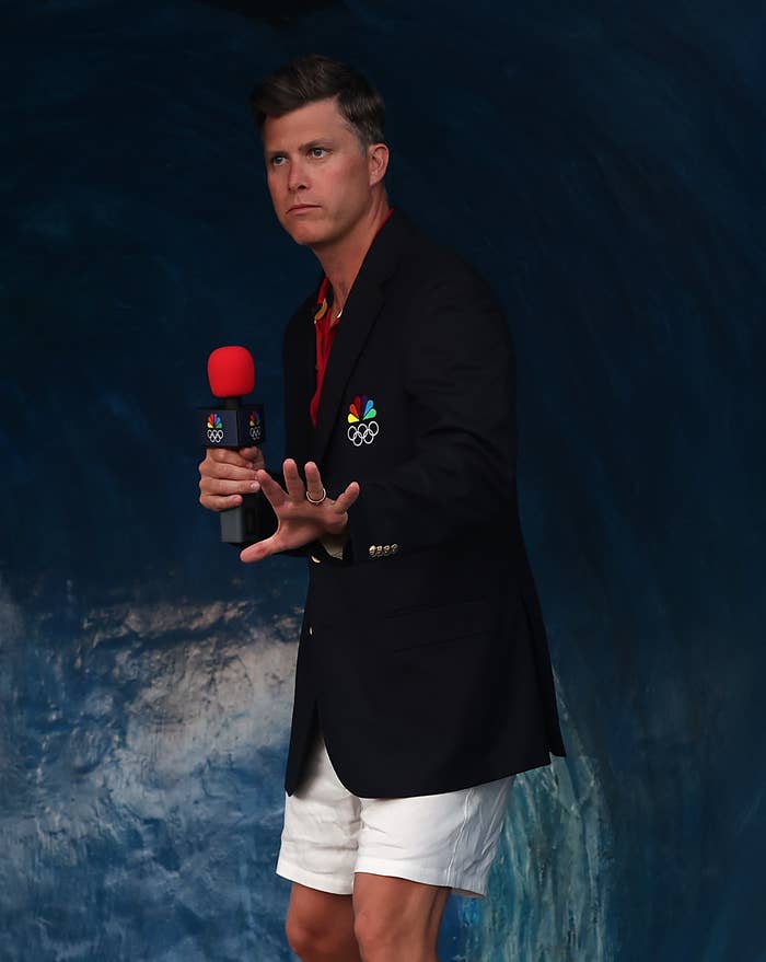Colin Jost wearing a blazer and shorts, holding a microphone, poses inside a blue tunnel at an event
