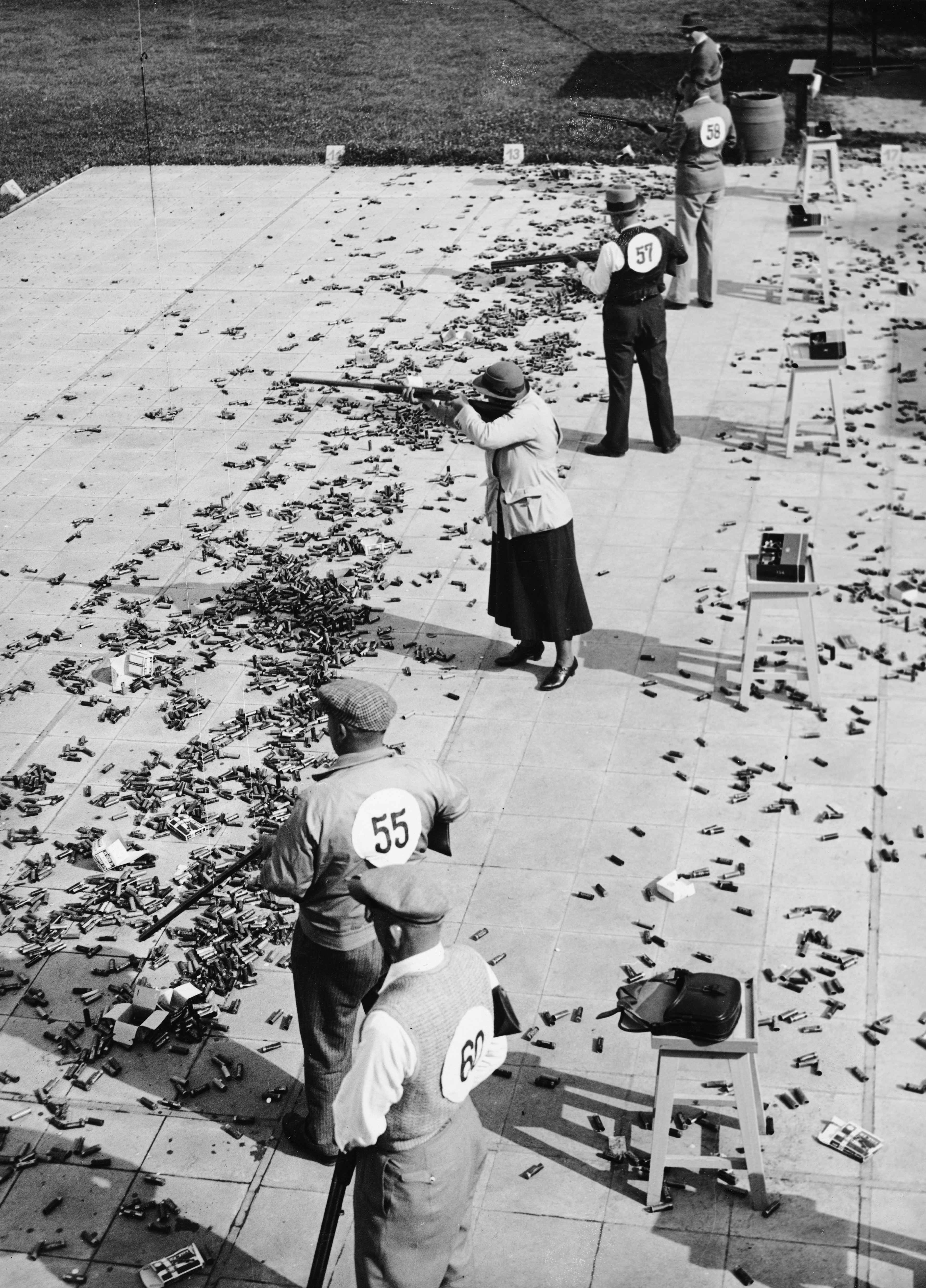 People with number tags (55, 57, 62) participate in a shooting competition, standing in line and aiming at targets. Spent cartridges cover the ground