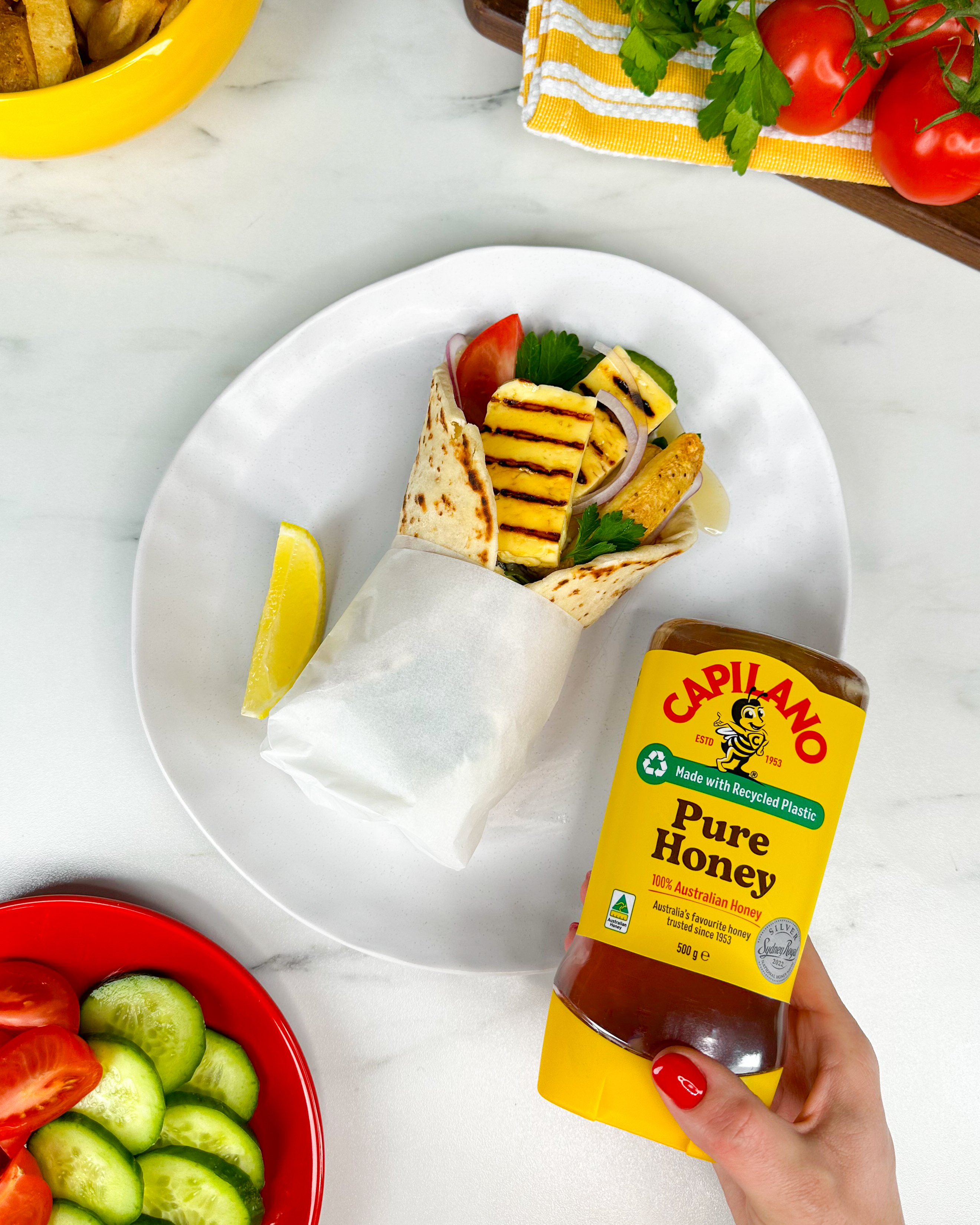 A hand holding a Capilano Pure Honey bottle over a wrap filled with grilled pineapple, vegetables, and a lemon wedge on a plate next to sliced vegetables