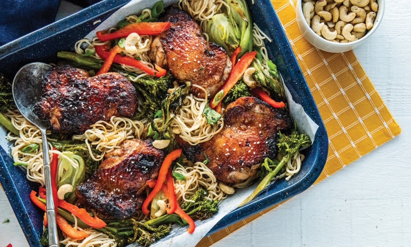 Close-up of a meal featuring roasted chicken thighs, noodles, red bell peppers, green vegetables, and a bowl of cashews on the side