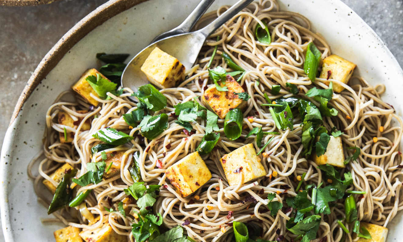 A bowl of noodle salad with grilled tofu cubes, chopped green vegetables, and garnished with fresh herbs, served with a fork and a spoon