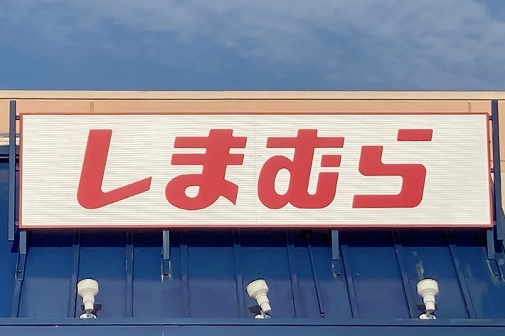 The image shows a storefront with Japanese text on a sign that reads &ldquo;しまむら&rdquo; above the entrance. There is also a handicap accessible sign visible in the lower right corner
