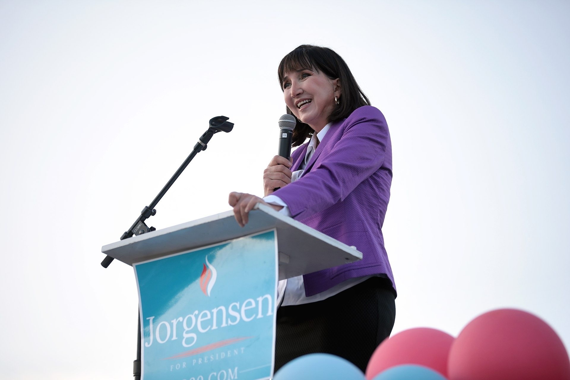 Jo Jorgensen speaks at a podium with a microphone. The podium has a campaign sign with her name