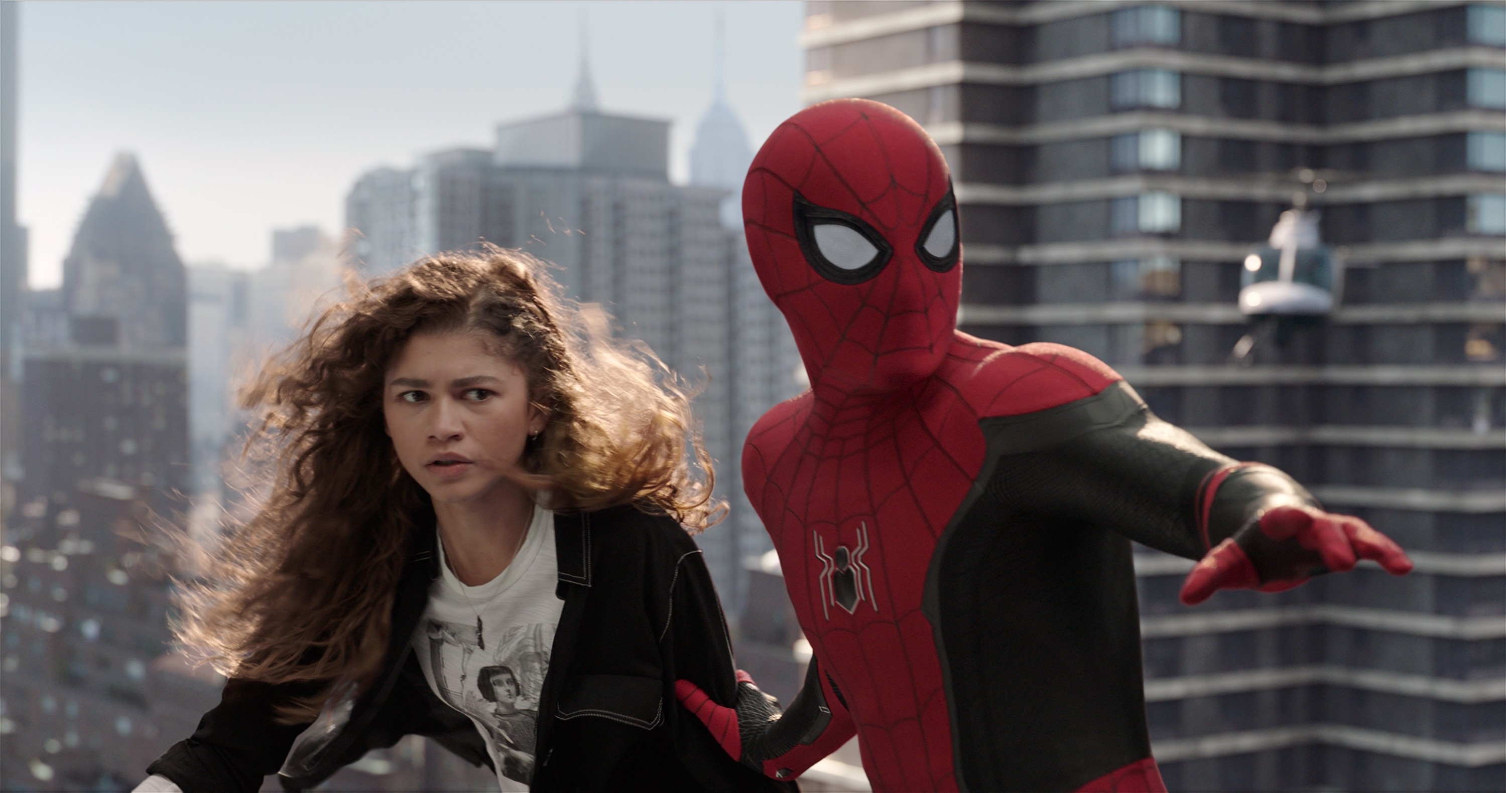 Zendaya and Spider-Man in an action scene with a cityscape background. Zendaya looks worried while Spider-Man extends his arm protectively