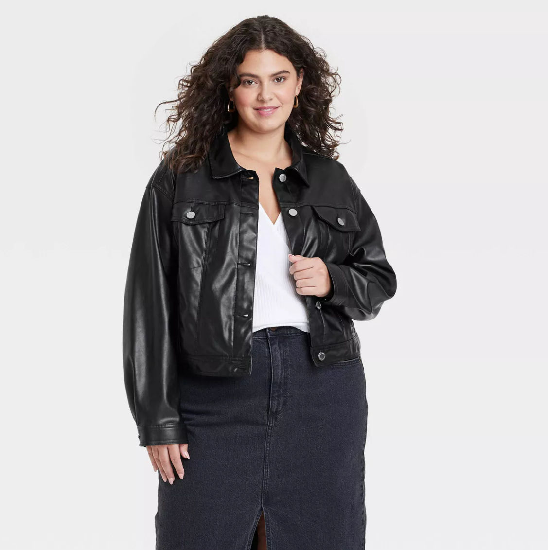 A model with curly hair wearing a black leather jacket, white top, and dark skirt poses, smiling against a plain background