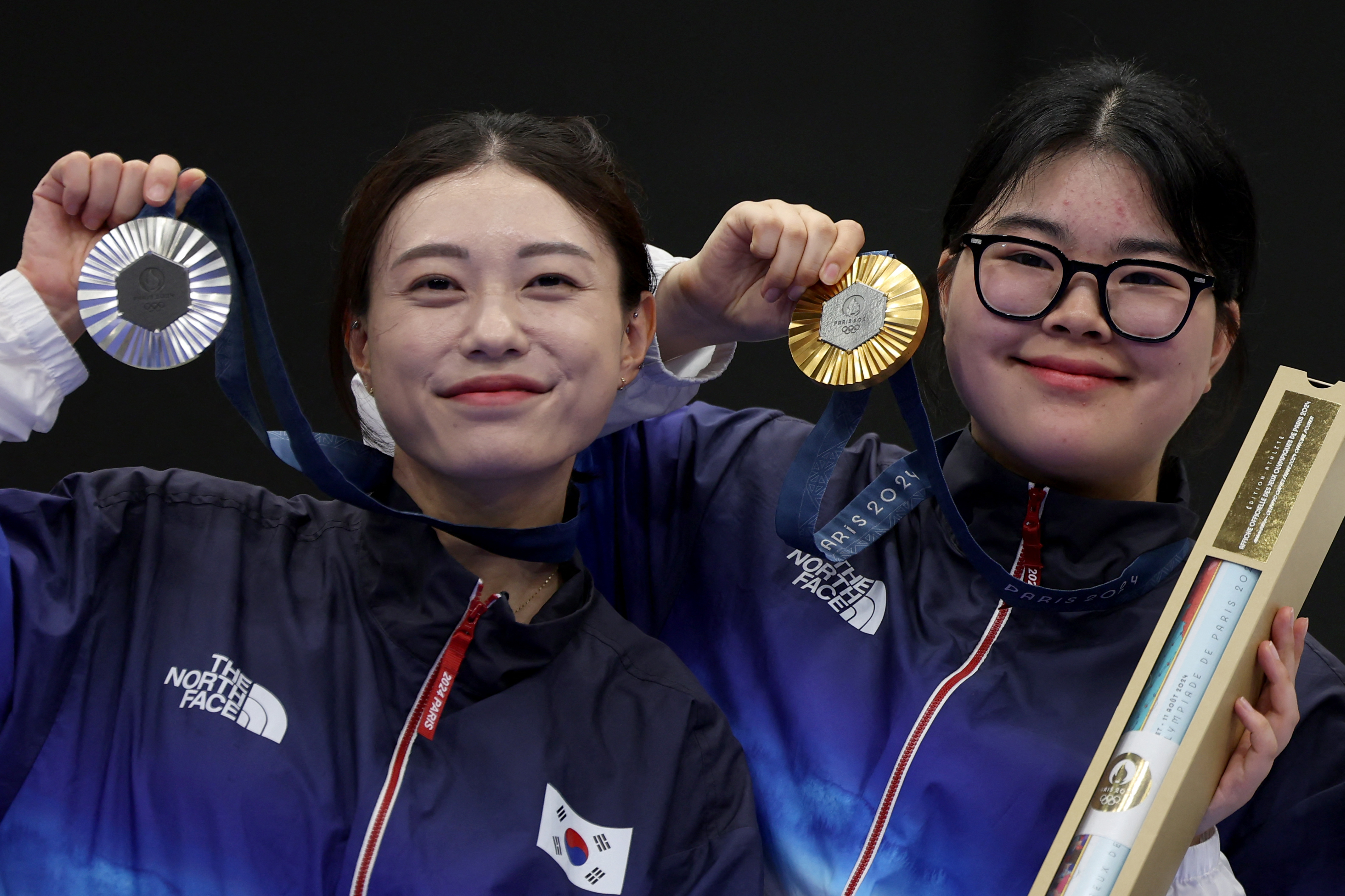 South Korean athletes Kim Ye-ji and Oh Ye-jin smile while holding up their Olympic medals and a certificate in their team jackets