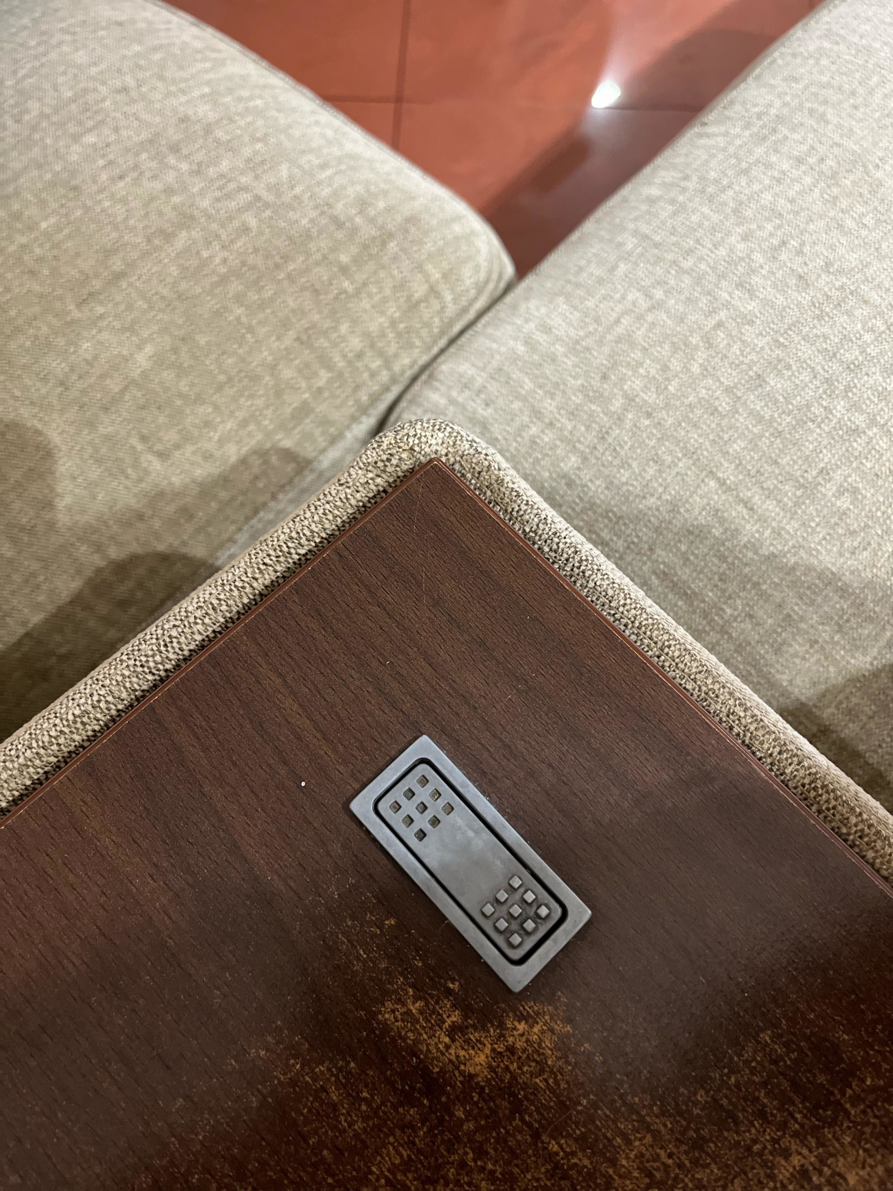 A small metallic remote control is embedded into the armrest of a beige couch next to a wooden surface
