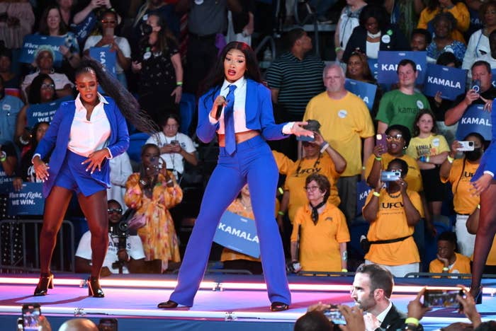 Megan Thee Stallion performs on stage wearing a vibrant blue suit beside a dancer at a Kamala Harris event. A diverse crowd is seen cheering in the background