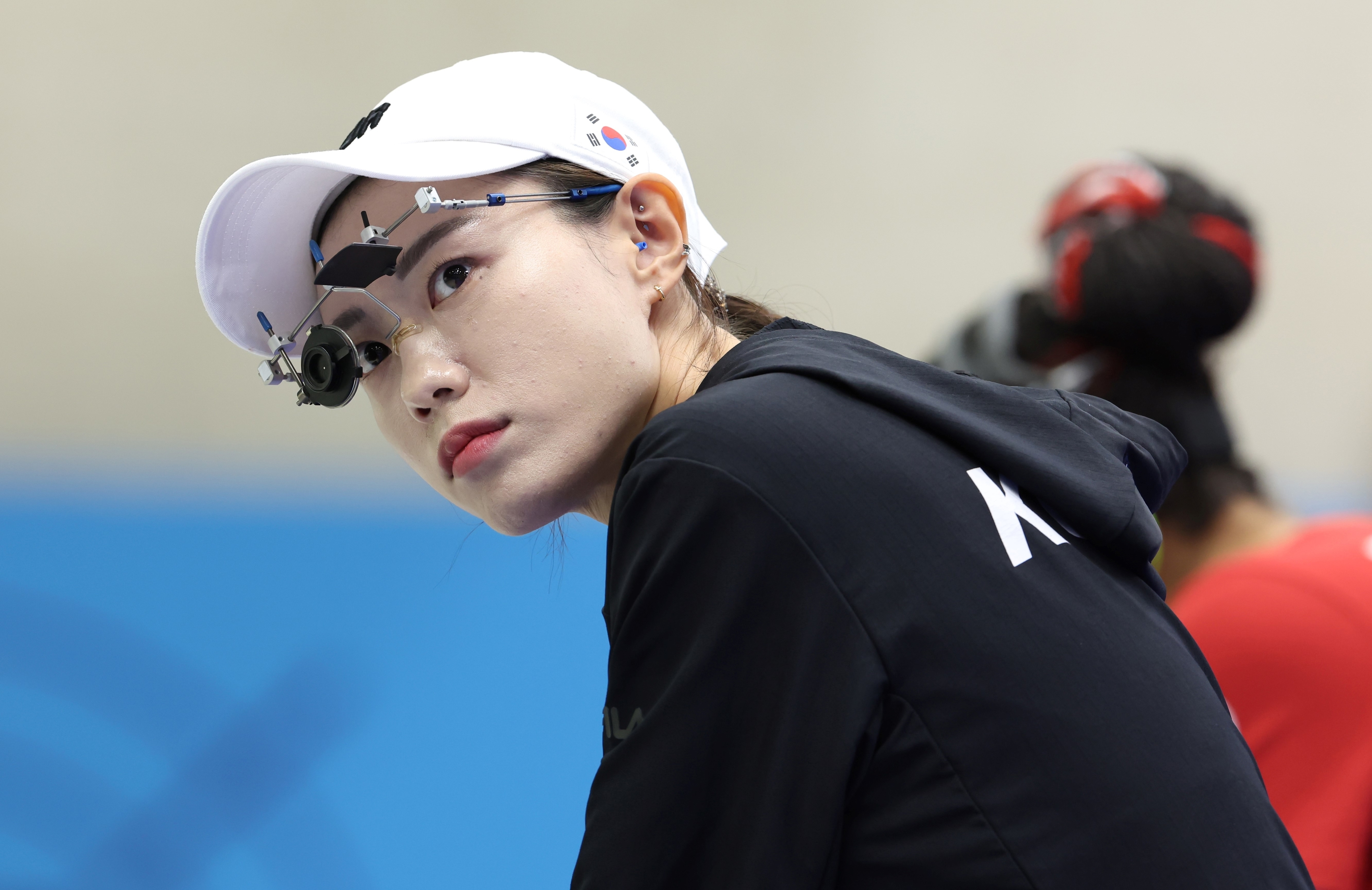Shooter Kim Ye-ji focuses intently during practice, wearing a white cap and precision eyeglasses. Another competitor is seen in the background