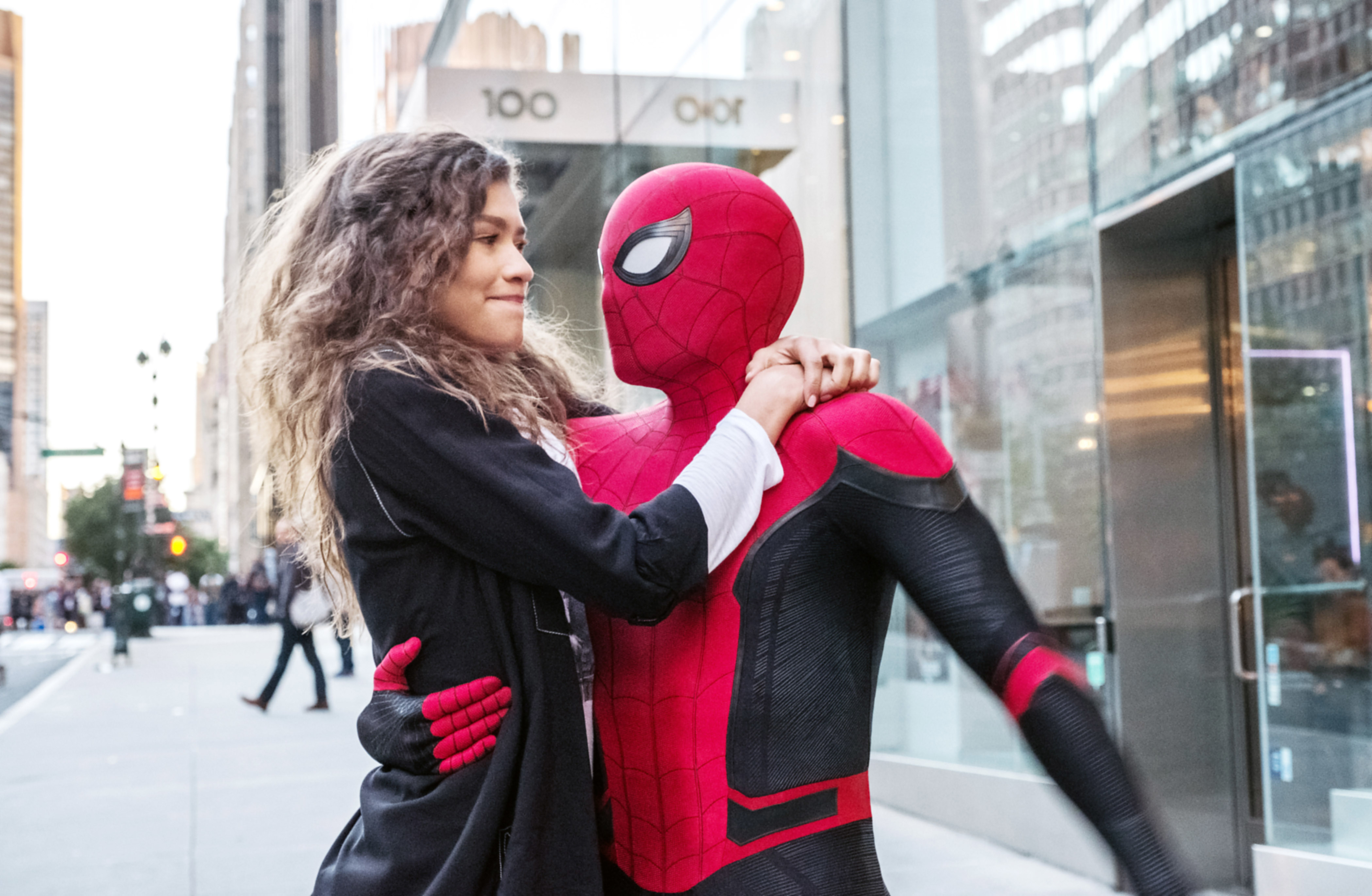 Zendaya embraces someone in a Spider-Man costume, posing on a city sidewalk in front of a glass building