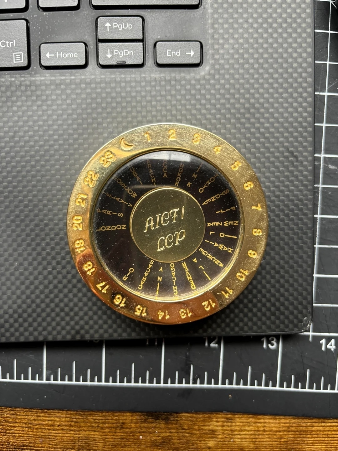 A brass device with the text "AICF1 LCP" in the center, placed on a textured surface next to a keyboard and ruler