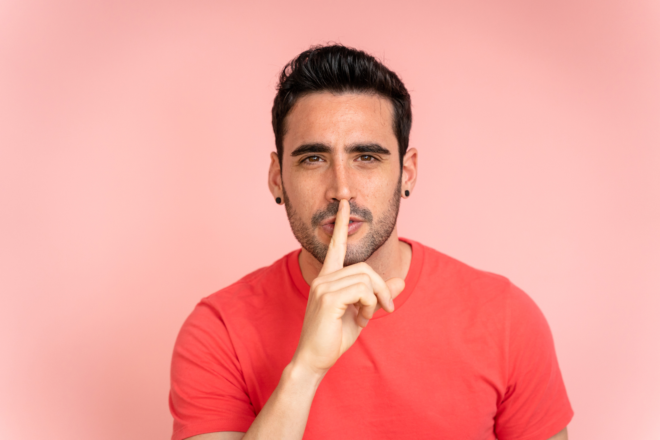 A man stands against a pink background, holding his index finger to his lips in a shushing gesture