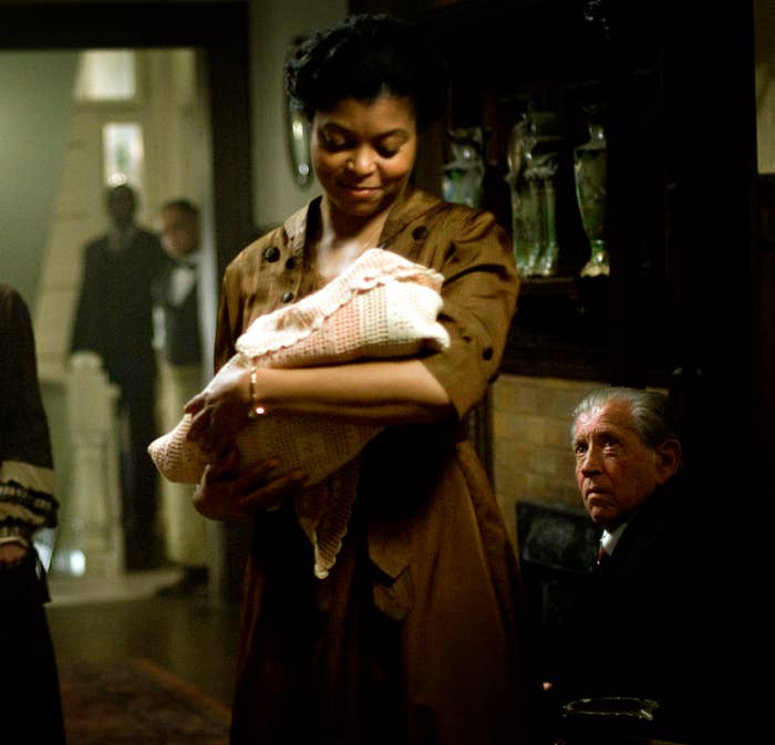 In a dim living room, six older people watch actress Taraji P. Henson, who is smiling and holding a swaddled baby